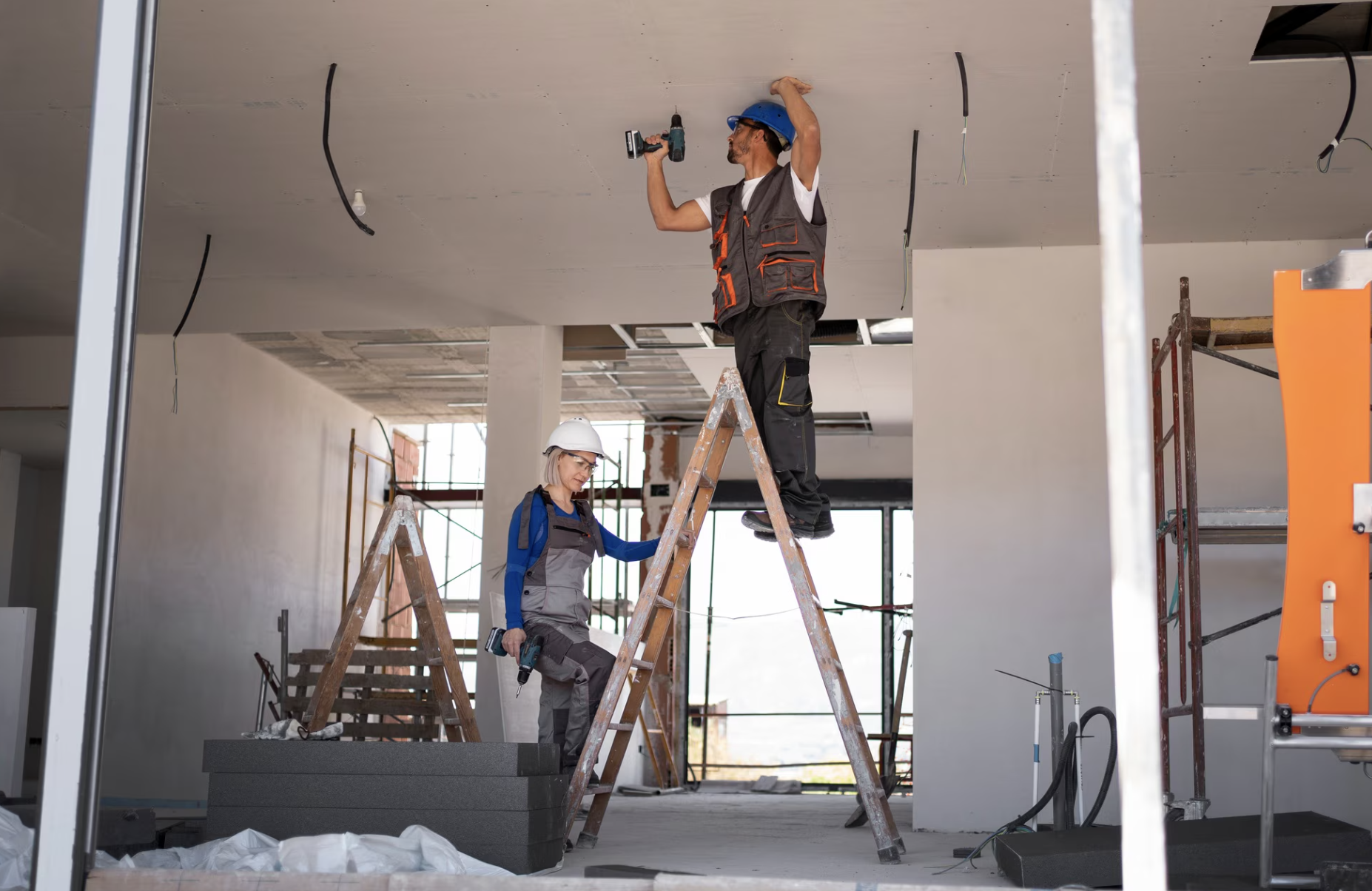 Deux ouvriers du bâtiment portant des casques de sécurité et des gilets de travail installent un plafond à l’aide de perceuses, l’un étant en haut d’une échelle et l’autre au sol, dans un intérieur en chantier.