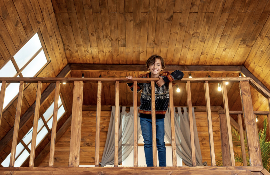 Enfant souriant s’appuyant sur une rambarde en bois dans une mezzanine chaleureuse sous un plafond mansardé en bois.