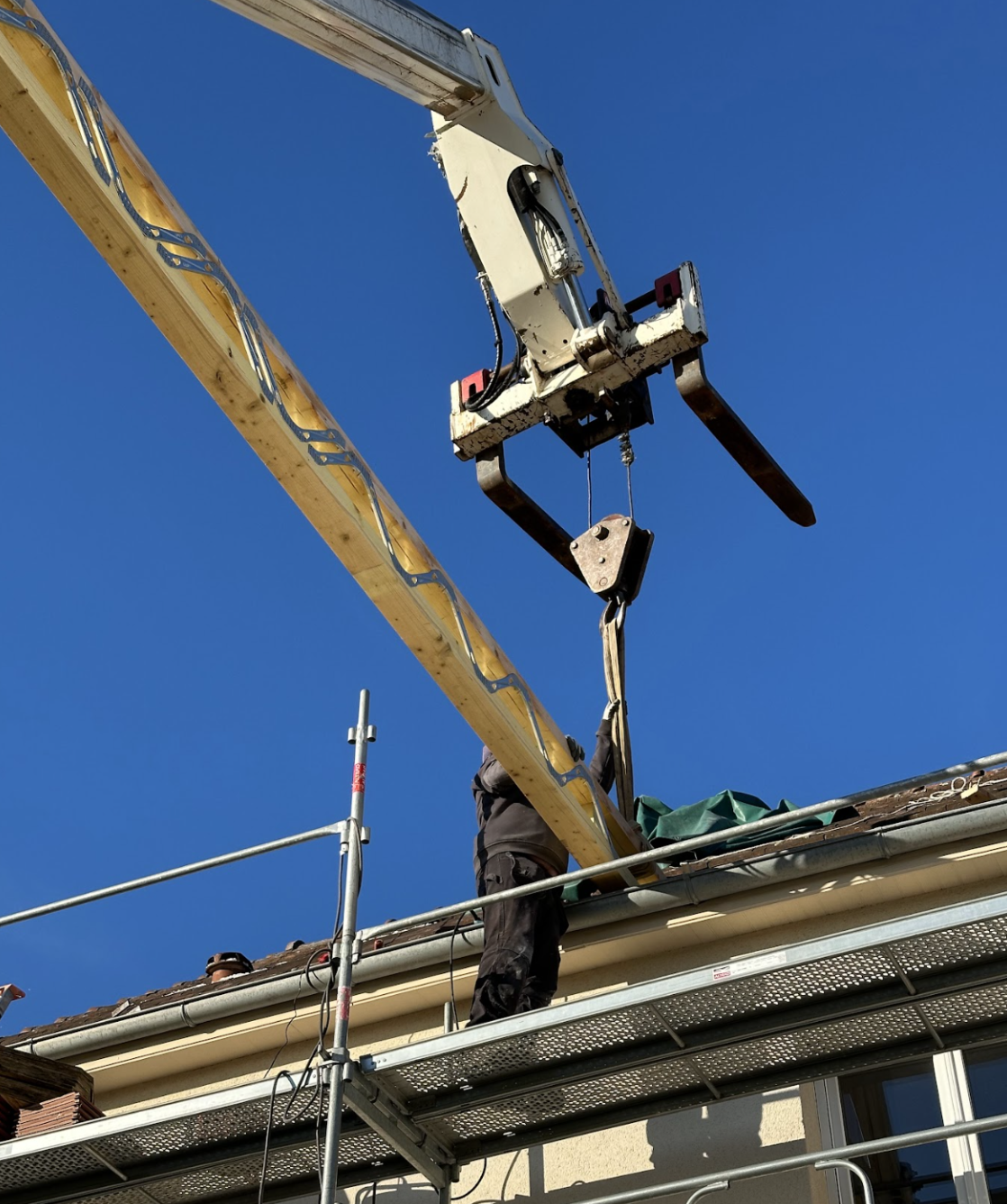 Opération de levage sur un chantier de toiture : un ouvrier sur un échafaudage guide une poutre en bois soulevée par une grue, sous un ciel bleu dégagé.